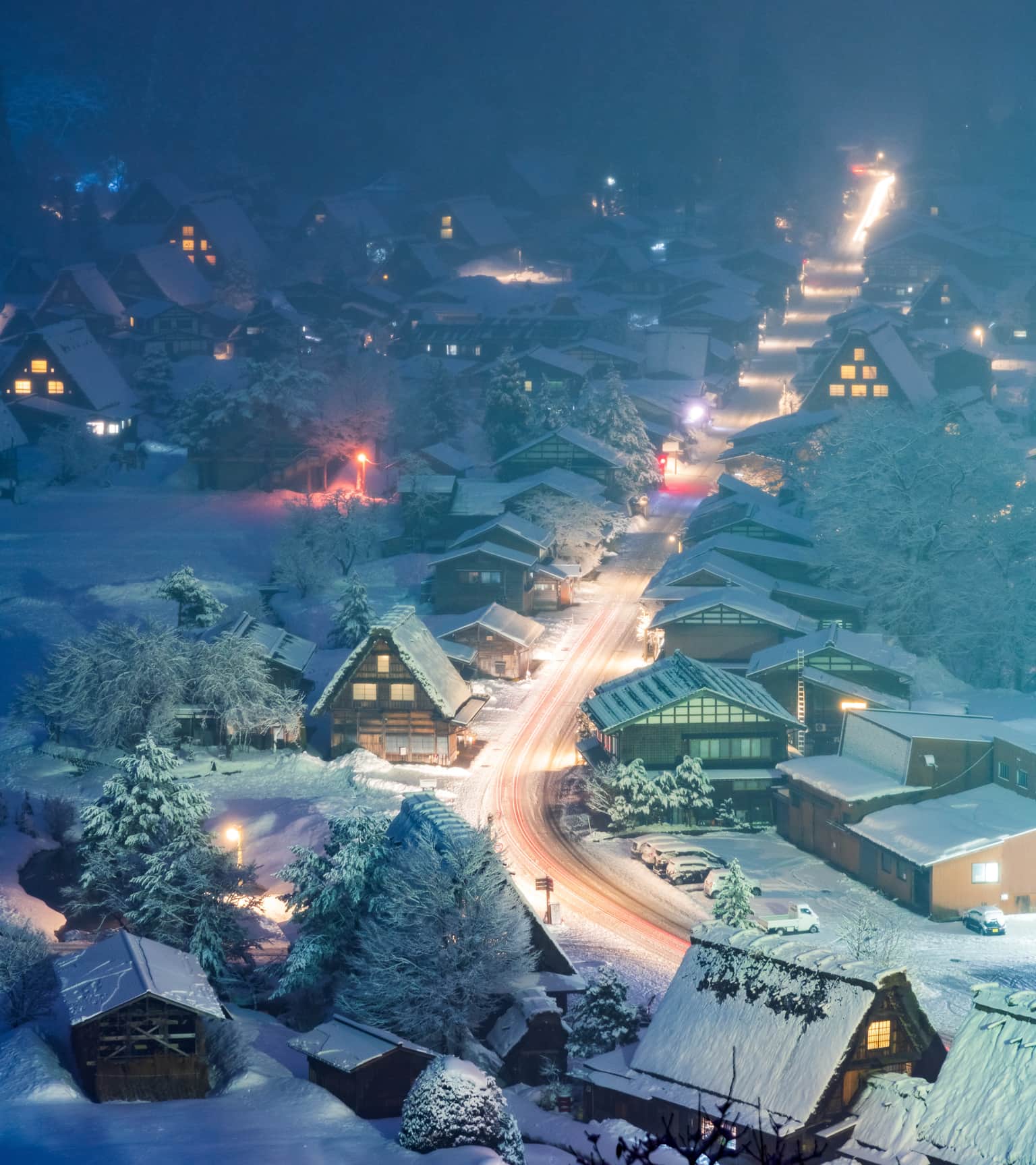 Snowy village at night with illuminated houses and winding road.