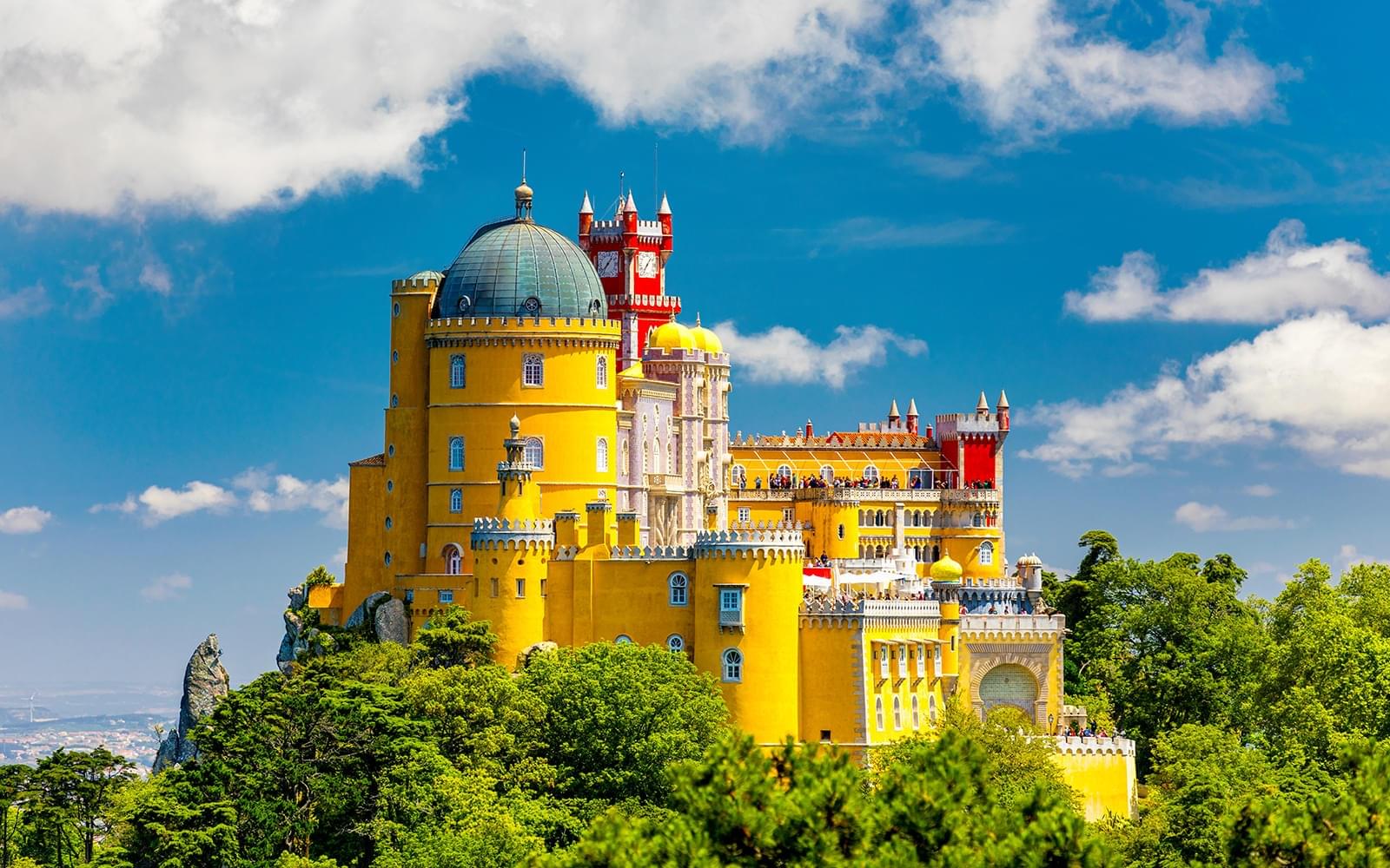 The vibrant yellow, red, and blue exterior of Pena Palace in Sintra, Portugal, standing majestically against a clear sky with lush green trees.